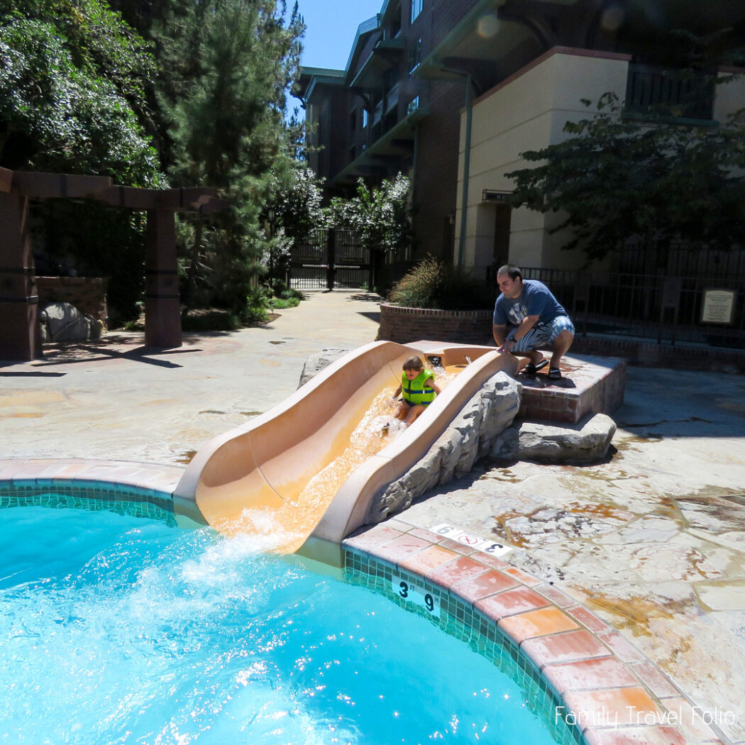 Dad helping toddler girl go down small water slide into pool.