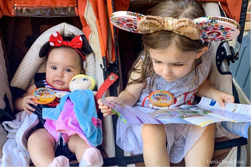 Toddler girl and baby sister sitting in a double stroller, wearing Minnie Mouse ears and 1st visit buttons. Toddler is looking at Disneyland map. 