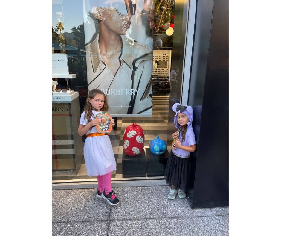 Two young girls in Halloween costumes standing next to Disney-themed pumpkins, including a Lilo and Stitch pumpkin, during Pluto's Pumpkin Pursuit in Downtown Disney.