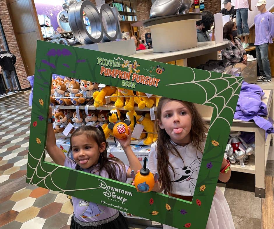 Two young girls holding mini pumpkin buckets and posing with a Pluto's Pumpkin Pursuit 2023 photo frame in World of Disney at Downtown Disney.