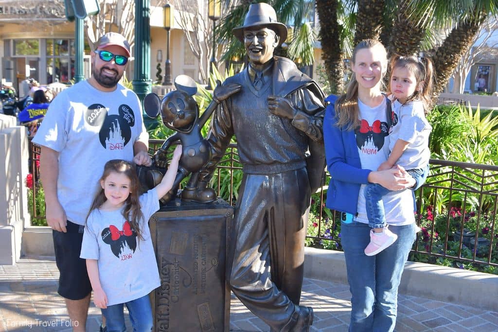 family of 4 wearing matching Disney Vacation t-shirts and standing next to Walt Disney with Mickey Mouse statue.