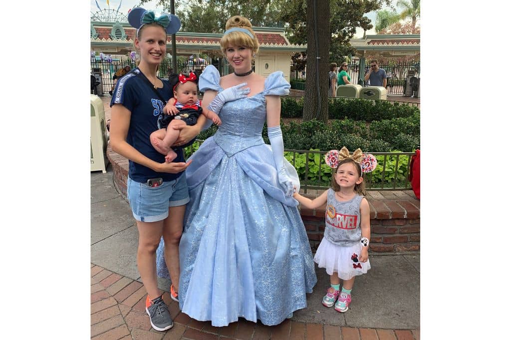 Mom holding baby girl with minnie mouse ears headband standing next to Cinderella with toddler girl in Marvel tank top and pink Minnie Mouse ears holds Cinderella's hand