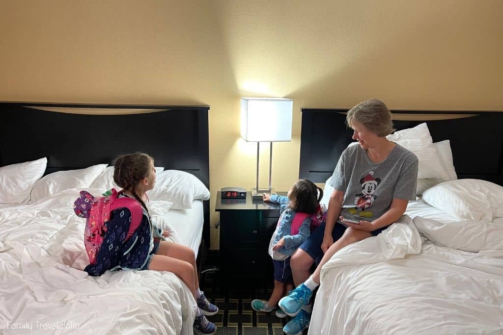 Grandma sitting on bed in hotel room with granddaughter sitting on the other bed and toddler granddaughter standing next to the bed pressing the button on the lamp.