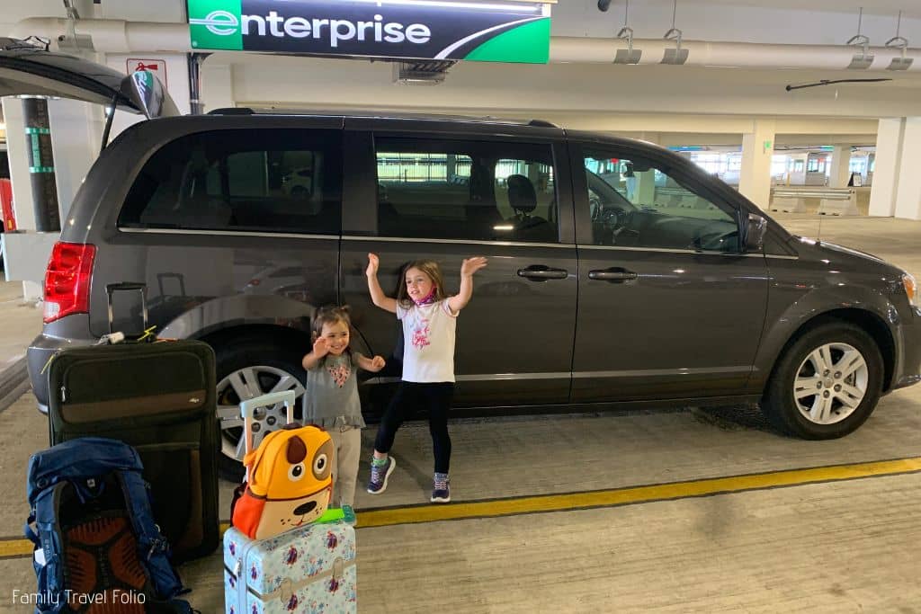 Little girl and toddler sister with arms up in front of rental car. Doggy backpack on top on Frozen roller carryon suitcase nearby.