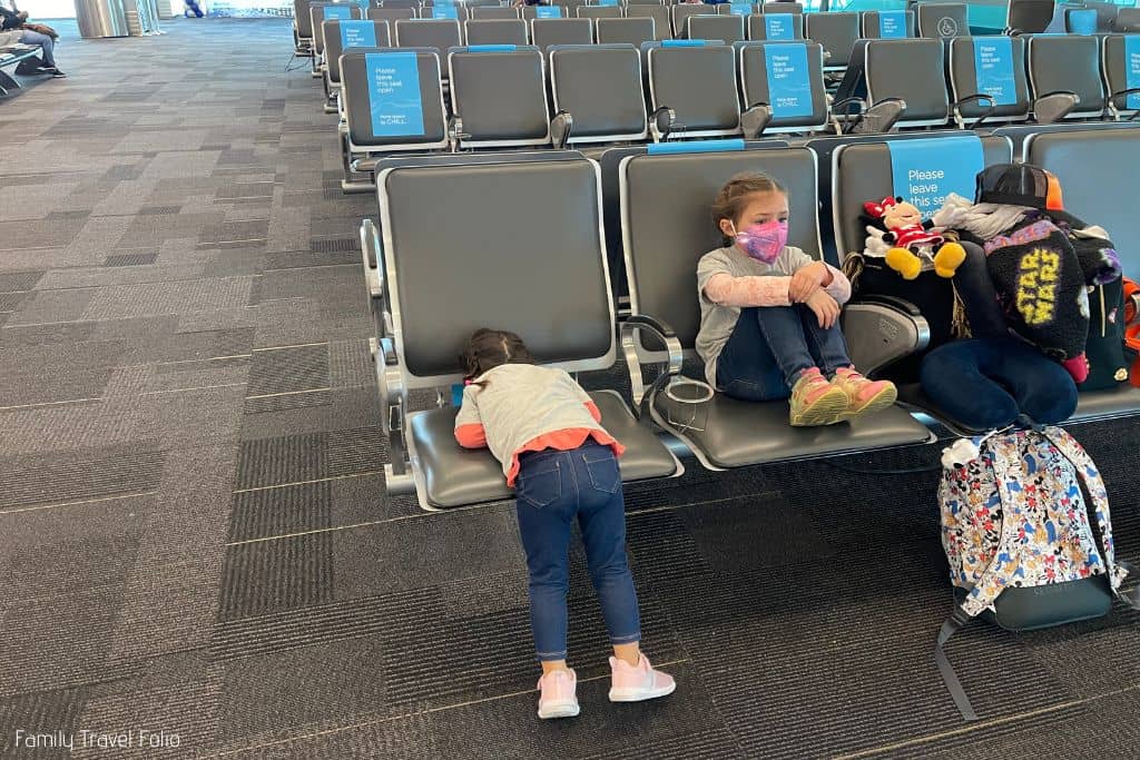 Little girl and toddler sister waiting on chairs at airport, luggage piled on seats next to them.
