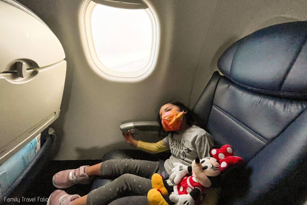 Toddler sitting in airplane seat with Minnie Mouse doll, airplane window open with light shining through.