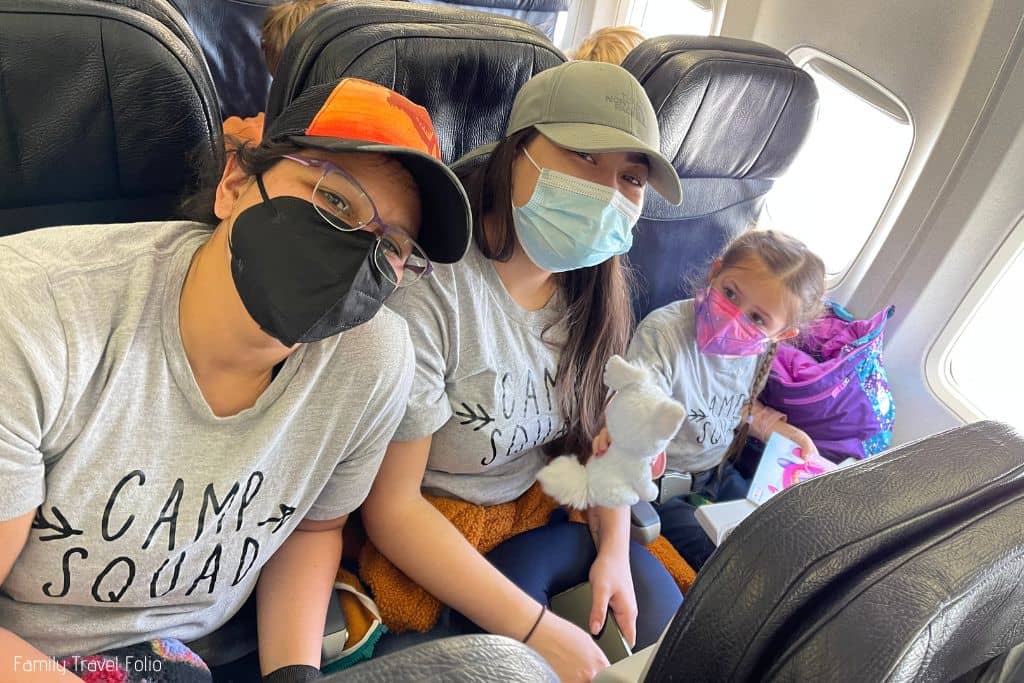 Two female friends and their niece sitting in a 3-seat airplane row. Niece is showing aunt her white stuffed animal dog.
