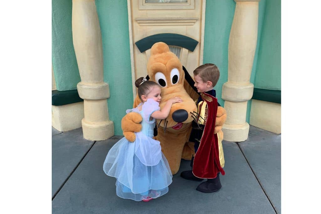 Toddler girl dressed as Cinderella and toddler boy dressed as knight giving Pluto a hug in ToonTown