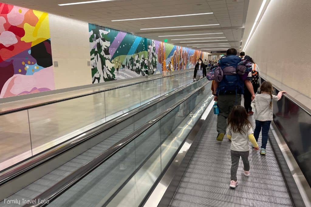 Dad walking on airport moving walkways with 2 young daughters following.