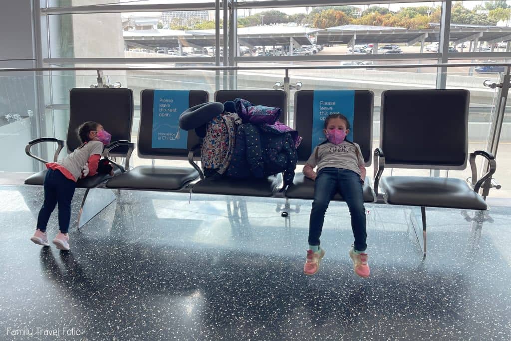 2 little girls waiting on chairs at airport. Luggage on seat in between them.