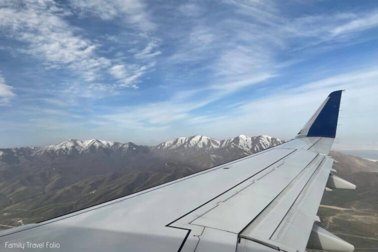View through airplane window of airplane wing and snowy mountains in distance.