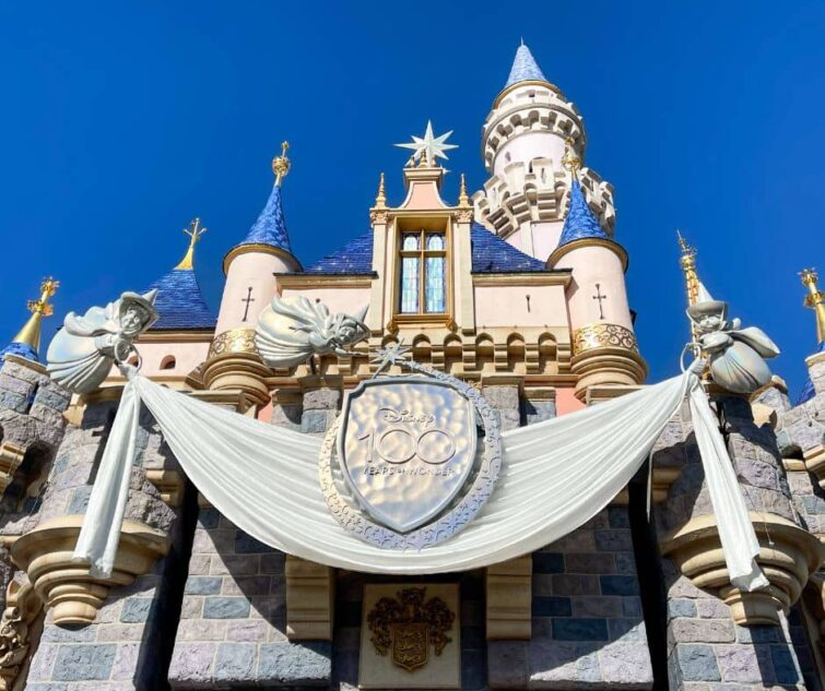 Disneyland castle with Disney 100 celebration platinum banner being held up by the 3 good fairies.
