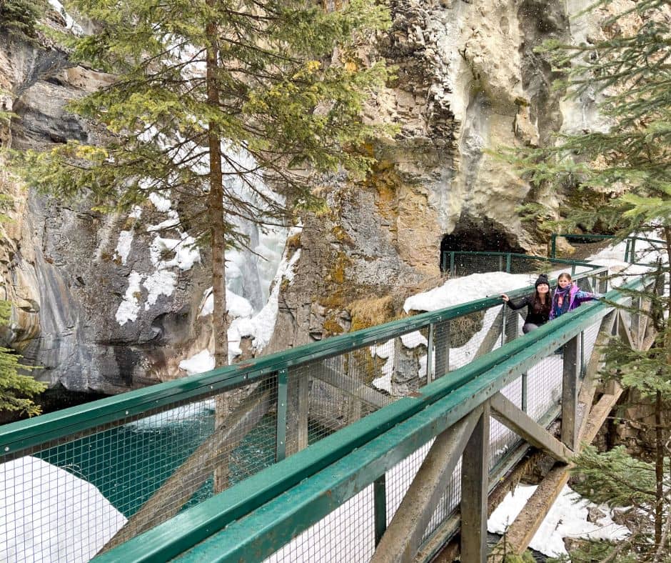 woman and little girl smiling on snow covered bridge to Johnston Canyon Lower Falls. behind them is the cave to the lower falls and the left side shows the waterfall behind them.