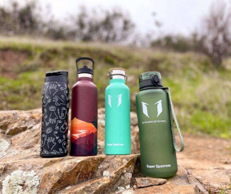 4 different Super Sparrow water bottles lines up in a row on a rock by a hiking trail.