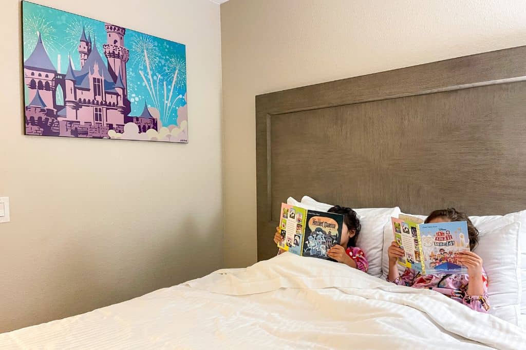 two girls in princess pajamas reading Disney books in Grand Legacy hotel bed.  A castle picture is on the wall behind them.