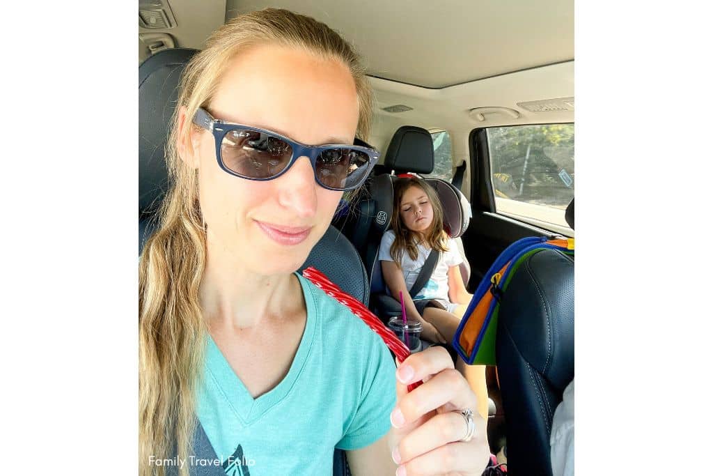Mother smiling with a candy Twizzler stick during a family road trip with her sleeping child in the backseat. Mom is smiling because she doesn't have to share the candy since the child is asleep in her car seat.
