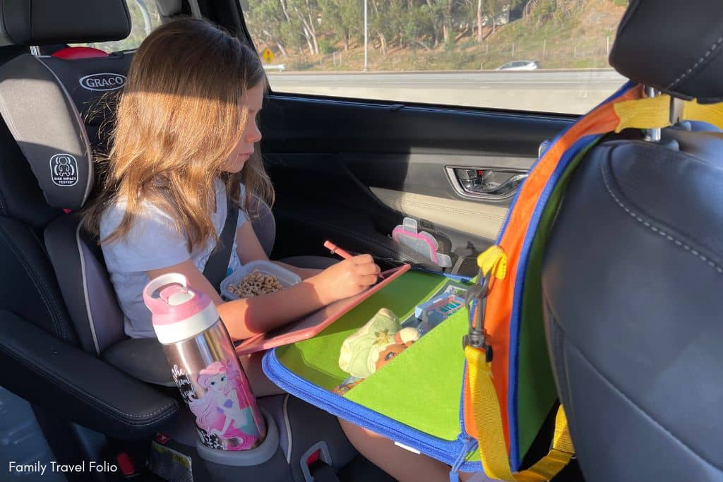 Child in a car seat focusing on a drawing pad with a bowl of Cheerios, a practical road trip snack for kids.