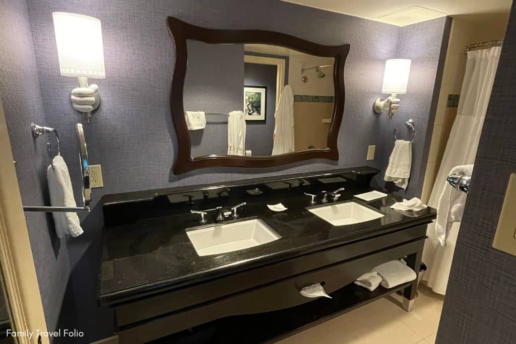 Elegant bathroom in the Disneyland Hotel featuring a large mirror and dual-sink marble vanity. Lights are held by Mickey Mouse white glove hands.