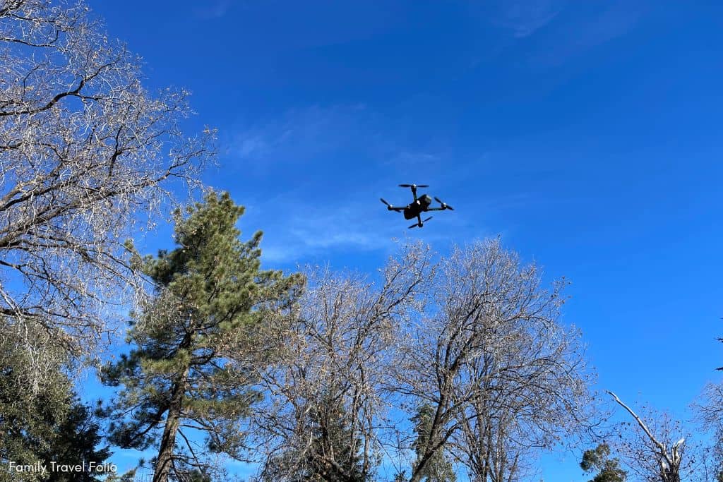 Black drone flying over green trees with blue sky
