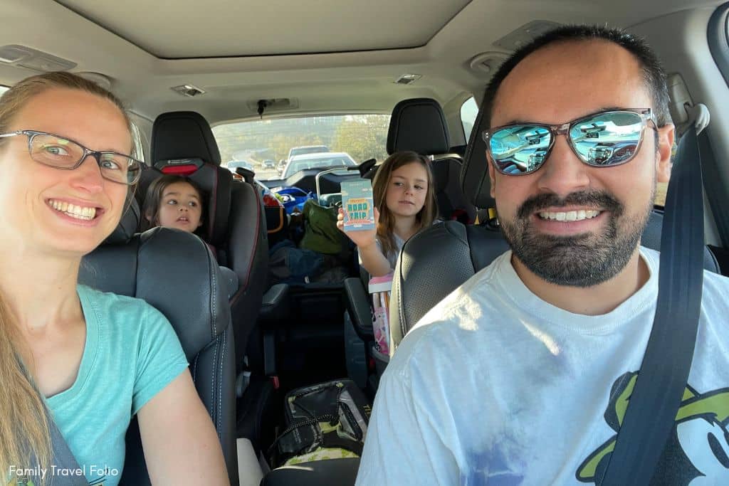Happy family on a road trip with two kids holding a book and a drawing tablet, enjoying each other’s company and ready with snacks for the journey.