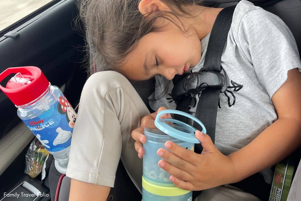 Sleeping child in a car seat clutching a stackable snack container, illustrating convenient access to road trip snacks for kids.
