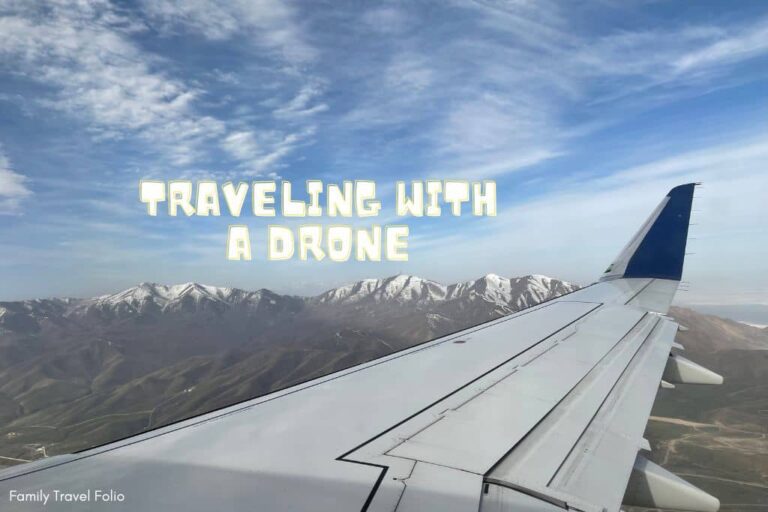 The words "traveling with a drone" in white block letters over blue sky with snow-capped mountain range in background and the wing of a plane in view.