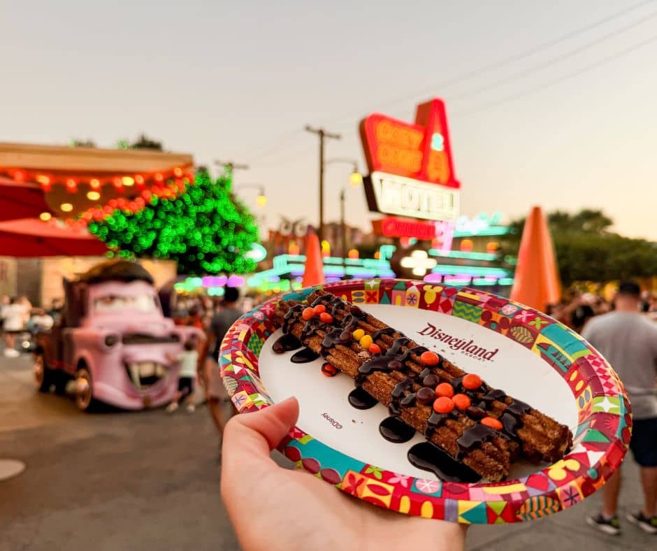 A chocolate-dipped churro covered in Halloween candy held up in Cars Land at Disney California Adventure, with Halloween lights and Mater in the background.