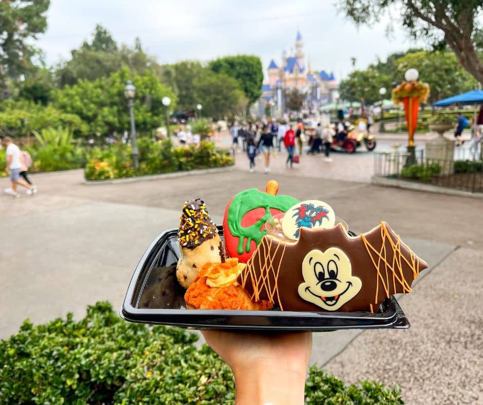 A tray of Halloween-themed treats at Disneyland, including a Mickey bat-shaped cookie, pumpkin cupcake, and poison apple caramel apple, with Sleeping Beauty Castle in the background during October.