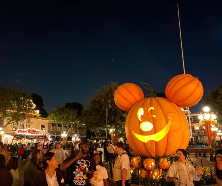 Giant Mickey Mouse pumpkin glowing brightly at night on Main Street, USA, surrounded by Disneyland visitors enjoying the Halloween atmosphere in October.