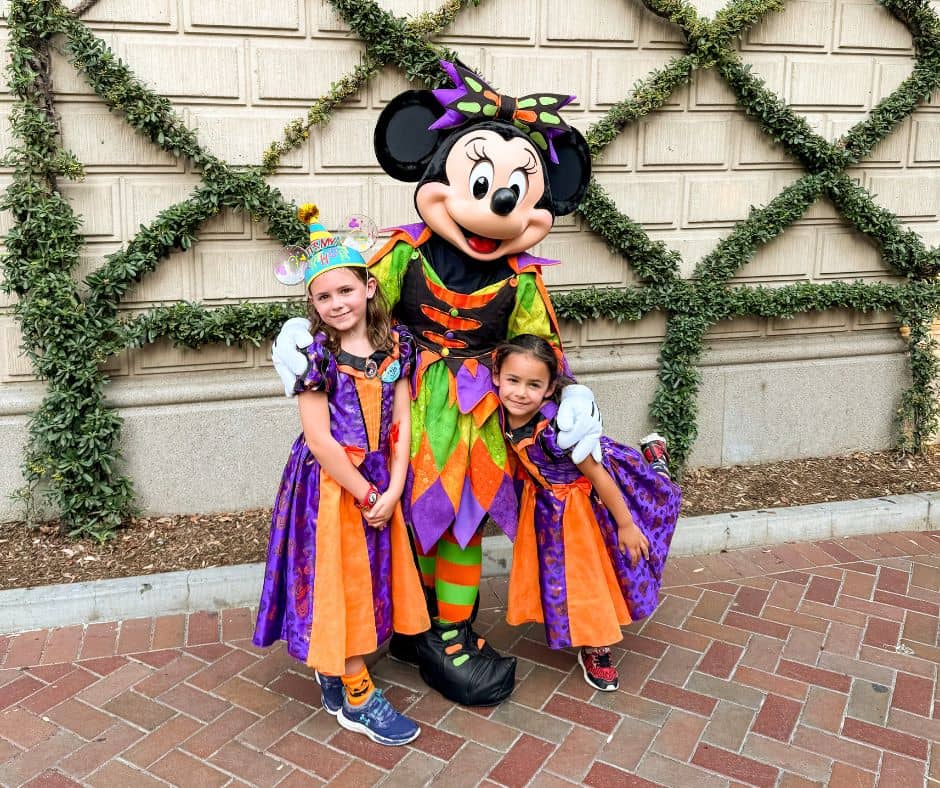 Two young girls in Halloween costumes posing with Minnie Mouse in her colorful Halloween outfit at Disney California Adventure during the 2024 Halloween celebration.