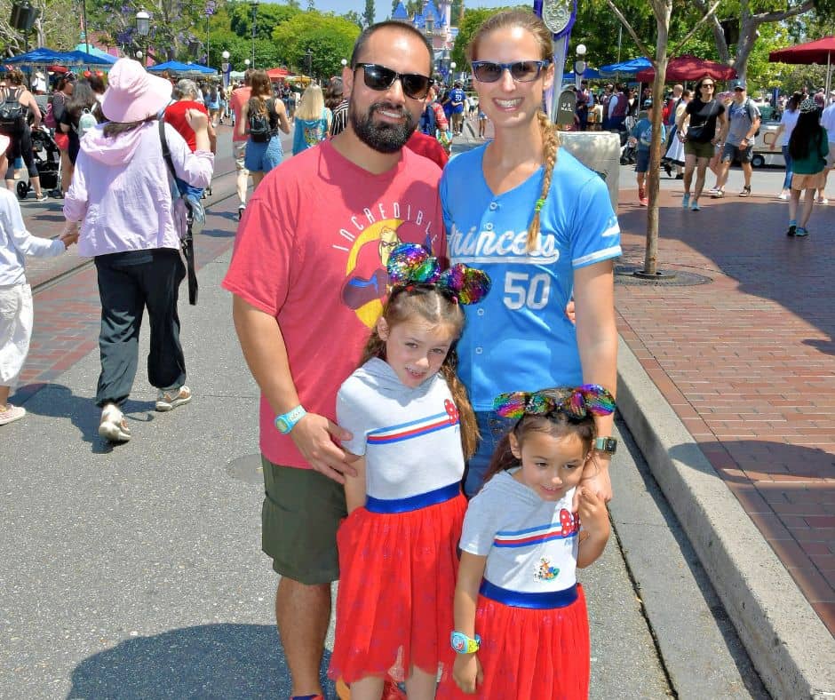 A family poses on Main Street at Disneyland in summer attire, wearing sunglasses and colorful shirts, prepared for hot weather at Disneyland.