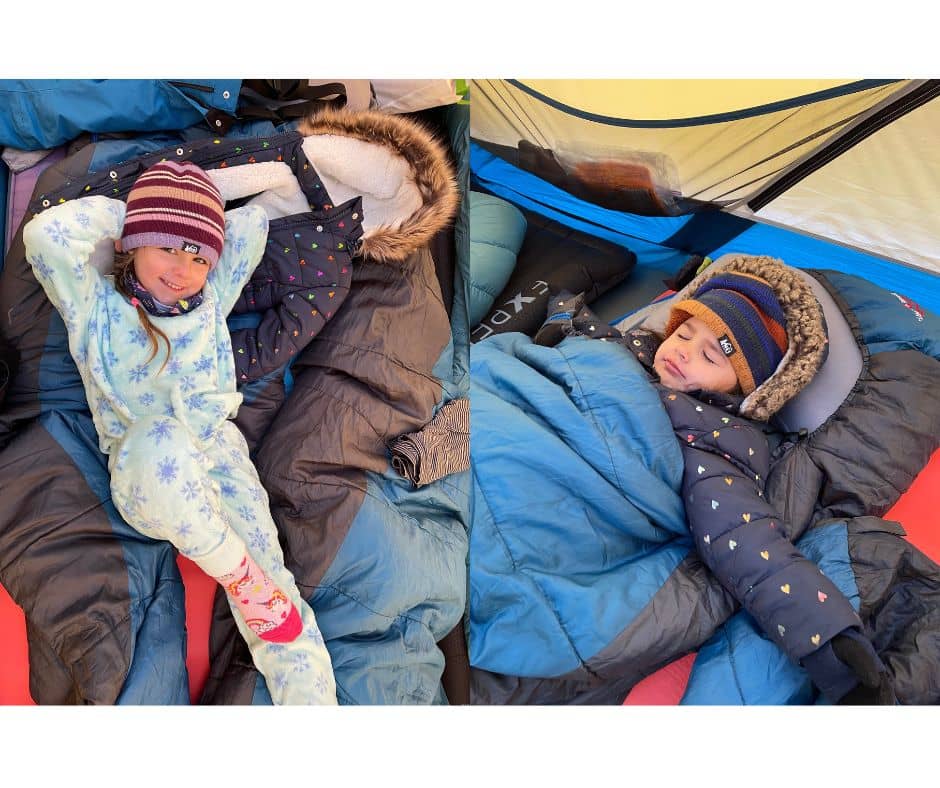 Two children bundled up in warm layers and sleeping bags inside a tent during a winter camping trip.