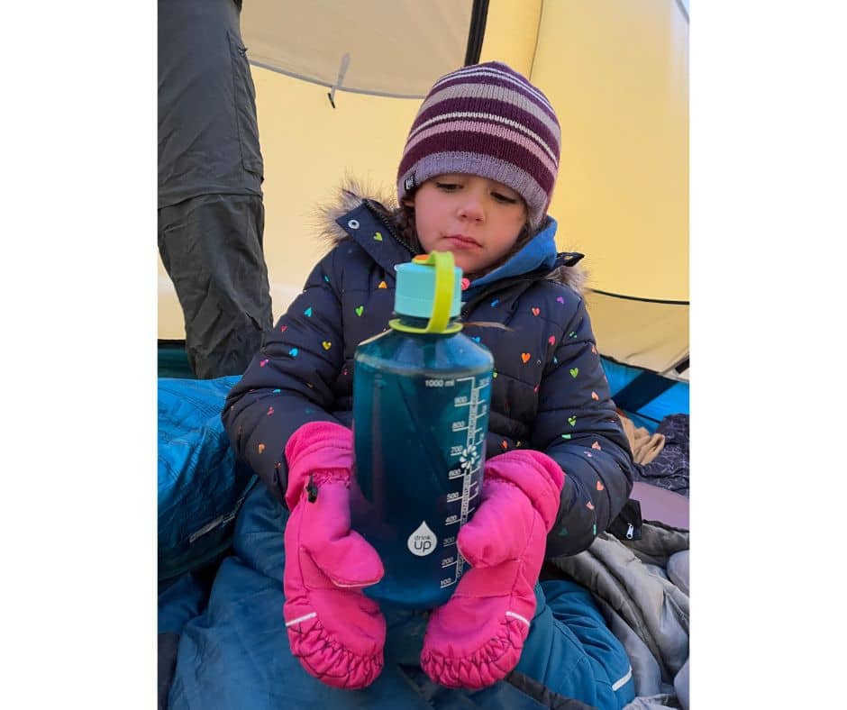 Child in warm winter clothing holding a water bottle inside a tent during a cold camping trip.