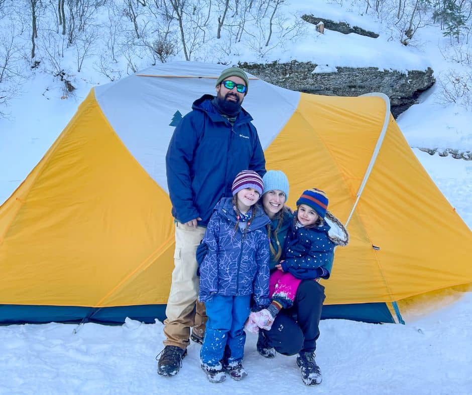 Family of four smiling in front of a yellow tent in the snow during a winter camping trip.