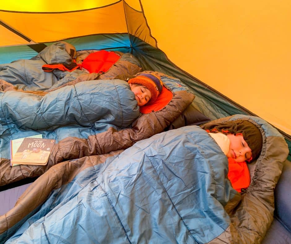 Two children snug in sleeping bags with liners inside a tent during a snowy camping adventure.