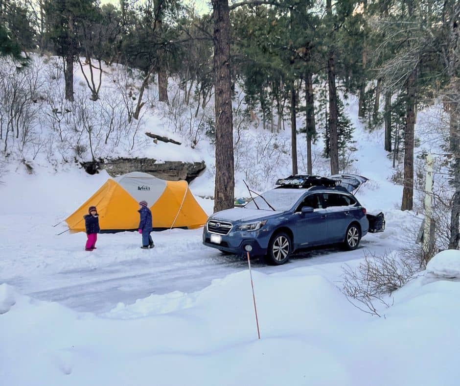 Snowy camping scene with a yellow tent and parked car, as children play in the snow.