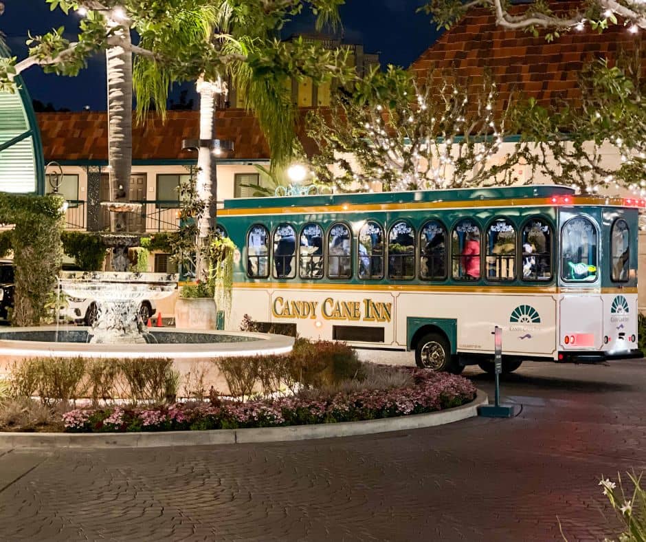 The Candy Cane Inn's signature green and white shuttle parked outside the hotel at night, surrounded by twinkling lights, a fountain, and lush greenery.