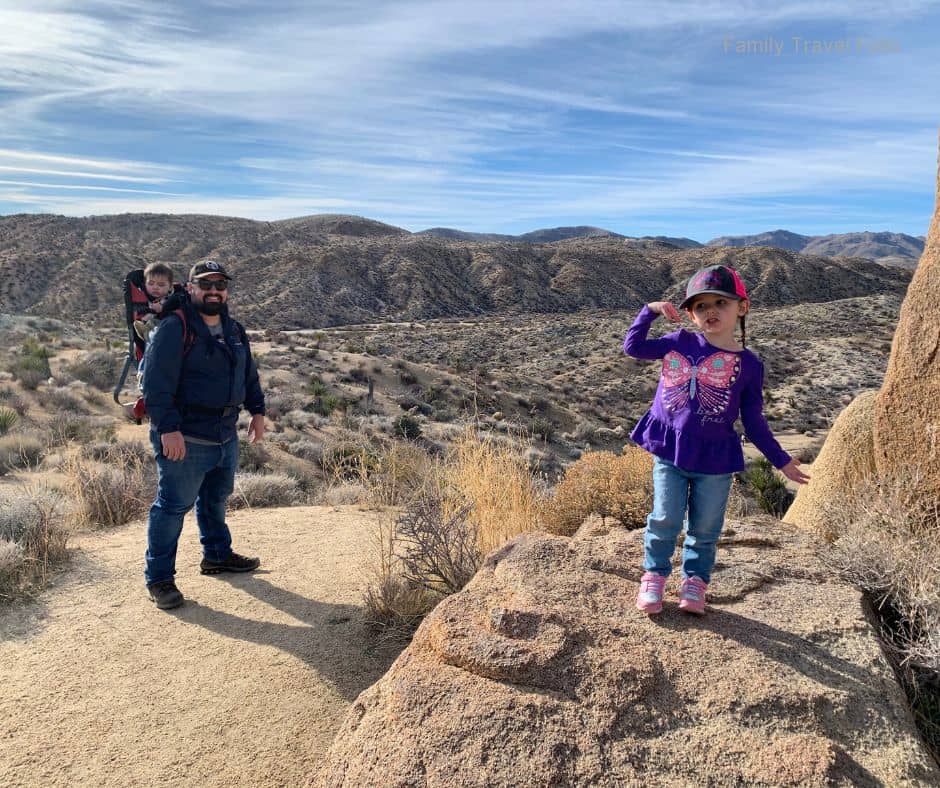 Dad hiking with two kids, one in a backpack carrier and the other posing on a rock along a desert trail