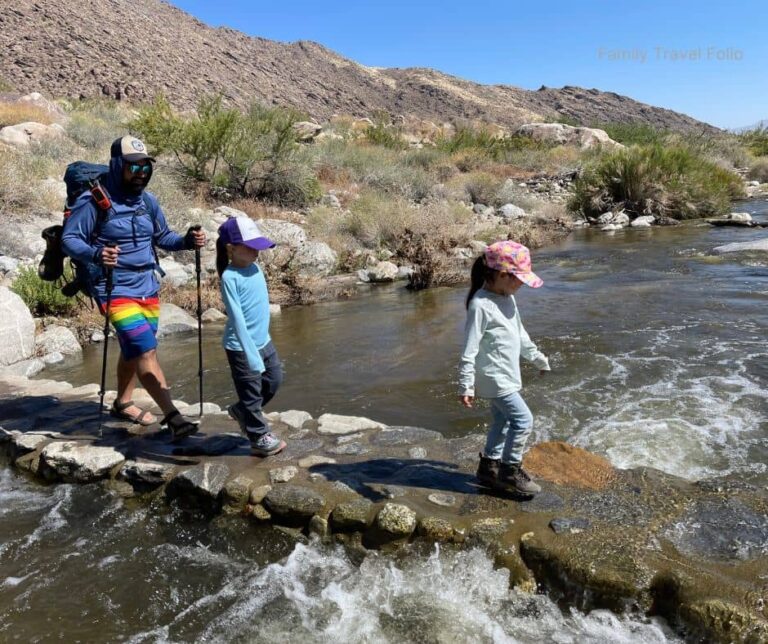 Dad and two kids crossing a rocky creek in hiking gear and sun hats on a family-friendly trail