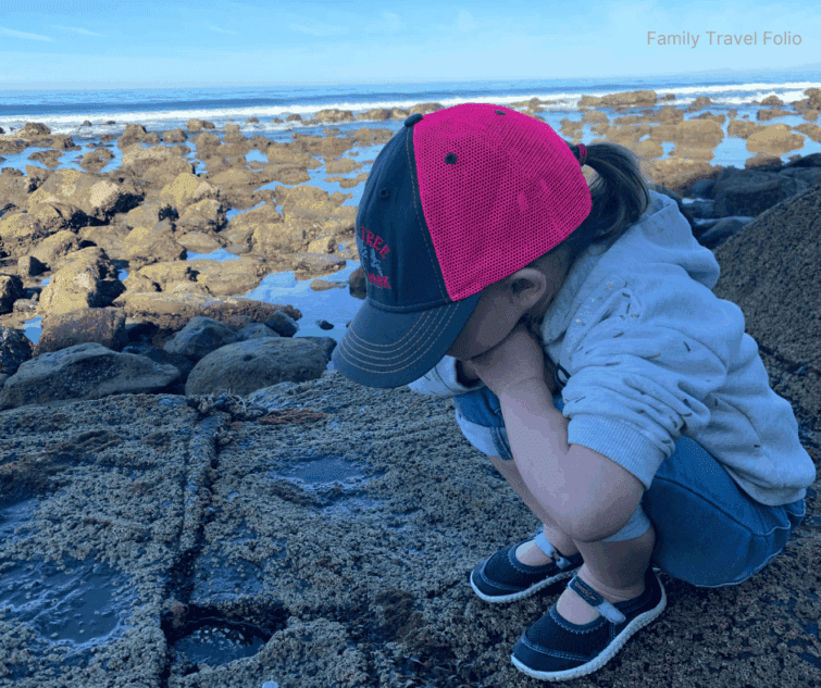 Child wearing a pink and black hat crouched on rocky tide pools by the ocean
