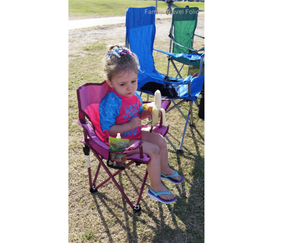 Young girl sitting in a pink camping chair eating a banana at an outdoor event with folding chairs nearby