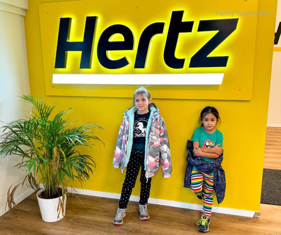 Two children standing by a bright yellow Hertz sign inside a Reykjavik car rental office, ready to start their Golden Circle road trip — one of the most convenient things to do in Reykjavik for families exploring Iceland.